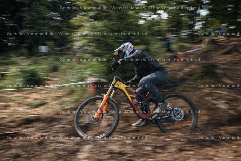 Downhill-Action | Das Bild zeigt einen Mountainbiker in voller Ausrüstung, der mit hoher Geschwindigkeit einen staubigen, unebenen Pfad in einem bewaldeten Gebiet hinunterfährt. Die Bewegung des Fahrers und des Fahrrads wird durch die lange Belichtungszeit dargestellt, was einen dynamischen Effekt erzeugt. Der Fahrer trägt einen Helm, eine Schutzbrille, Handschuhe und einen schwarzen Anzug. Das Fahrrad ist ein Downhill-Mountainbike mit roten Akzenten und einer robusten Federgabel. Der Hintergrund besteht aus dichtem Grünzeug und Bäumen. - Realisiert mit Pictrs.com