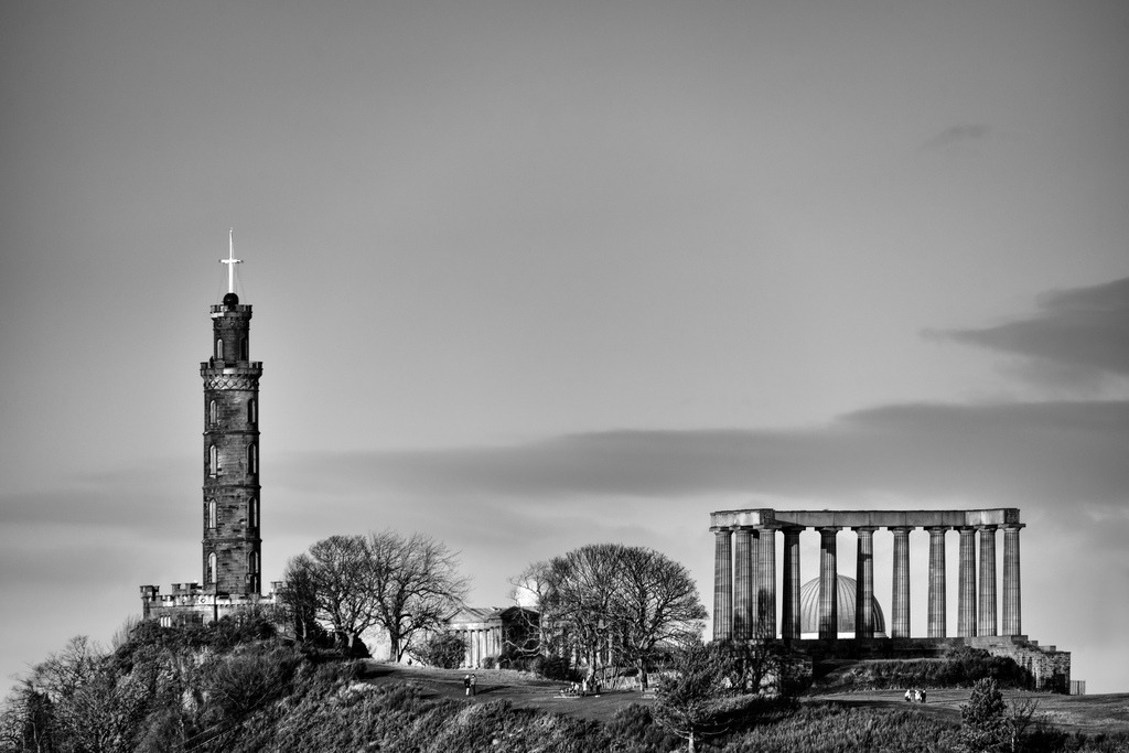Historische Monumente Calton Hill Edinburgh | Das Nelson Monument und das unvollendete National Monument dominieren die Skyline von Calton Hill in Edinburgh. Die Schwarz-Weiss-Aufnahme betont die architektonischen Details und die dramatische Form der historischen Bauwerke. Die Komposition mit weitem Himmel und kahlen Baeumen vermittelt eine zeitlose, ruhige Stimmung. - Realisiert mit Pictrs.com