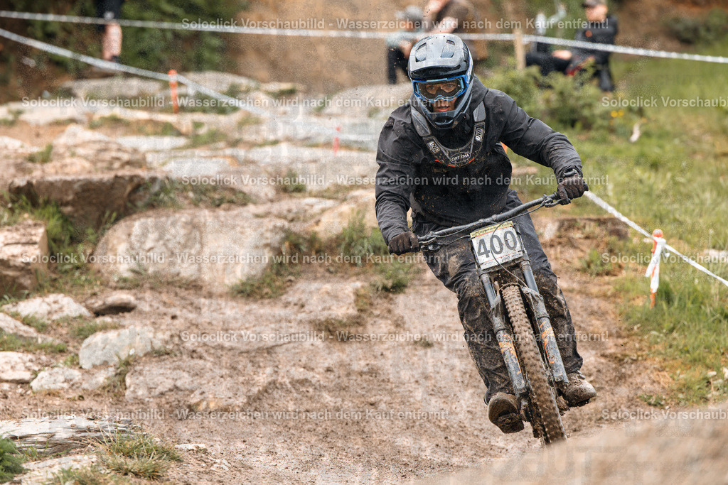 Mountainbike-Action im Schnee | Ein Mountainbiker in voller Ausrüstung rast einen staubigen, unebenen Pfad hinunter. Er trägt einen schwarzen Helm mit einer schwarzen Visier, eine dunkle Brille, eine schwarze Jacke und Handschuhe. Auf seinem Rücken befindet sich eine Nummer 400. Der Hintergrund zeigt eine verschneite Berglandschaft mit Bäumen und Felsen. Der Pfad ist mit Staub bedeckt, was darauf hindeutet, dass er gerade benutzt wurde. Der Fahrer scheint sich auf einen Sprung oder eine Abfahrt vorzubereiten. - Realisiert mit Pictrs.com