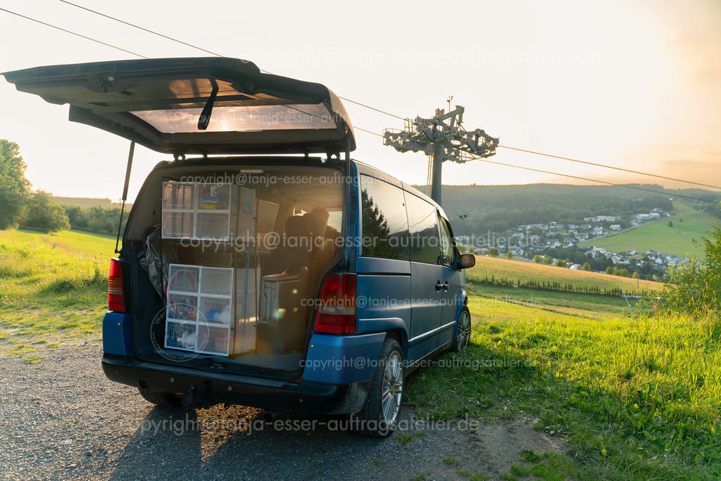 Auf dem Ettelsberg in Willingen parkt ein Van mit eingebautem Schrank | Vanlife. Blauer Van parkt vor einer schönen Aussicht in Willingen Upland in Deutschland. Neben dem Fahrzeug ist ein Skilift. Der Kofferraum ist geöffnet. Selbstgebautes Regal mit Haushalts Utensilien. Sonnenuntergang.