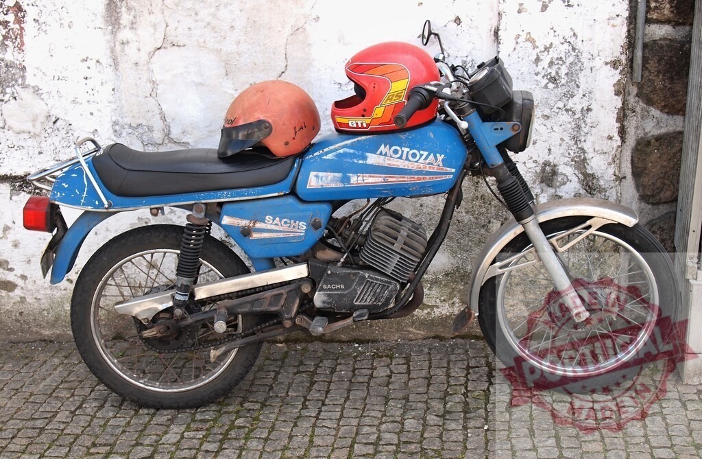 Old Motozax Motorcycle Parked near White Wall with Two Helmets | Old, slightly rusty blue Motozax motorcycle with two helmets parked near a weathered white wall - Realisiert mit Pictrs.com