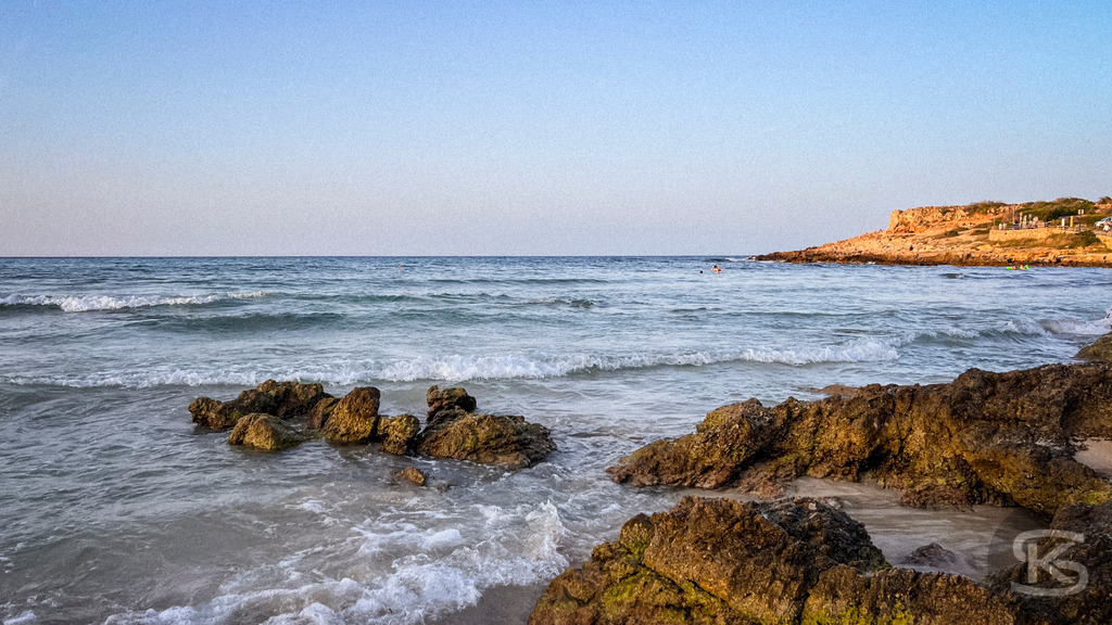 Abendruhe an der Cala Agulla – Exklusives Wandbild der mallorquinischen Felsenküste | Nahaufnahme von dunklen Küstenfelsen im ruhigen Wasser der Cala Agulla auf Mallorca während der Abenddämmerung mit Blick auf die entfernte Silhouette der Küstenlinie. - Realisiert mit Pictrs.com