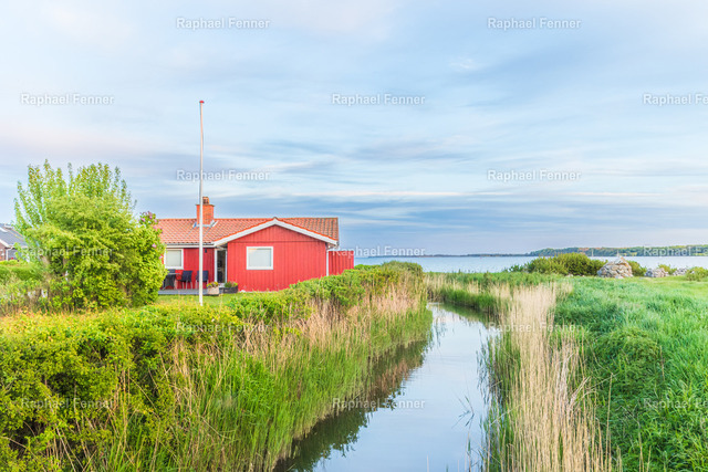 Rotes Ferienhaus an der dänischen Küste | Ein typisches rotes Ferienhaus eingebettet in die ruhige Küstenlandschaft Dänemarks. Umgeben von Schilf, einem kleinen Wasserlauf und der Weite des Meeres vermittelt dieses Bild die entspannte Atmosphäre eines nordischen Sommertages. Die harmonischen Farben und die natürliche Umgebung machen dieses Motiv zu einem zeitlosen Blickfang für alle, die Skandinavien und seine besondere Gelassenheit lieben.Als hochwertiger Fine-Art-Druck bringt dieses Motiv die Ruhe und Schönheit der dänischen Küste in Wohnräume, Ferienhäuser oder Büros.
