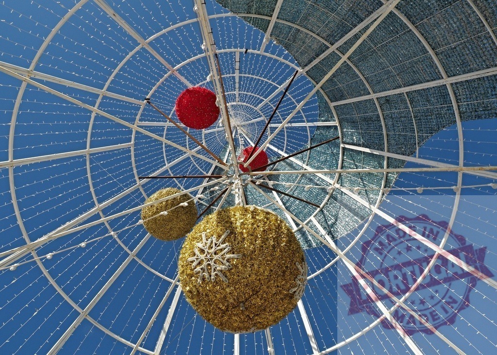Christmas decorations hanging from a large metal christmas tree structure with lights and blue sky background | Looking up at christmas decorations hanging from a large metal christmas tree structure with lights and blue sky background - Realisiert mit Pictrs.com
