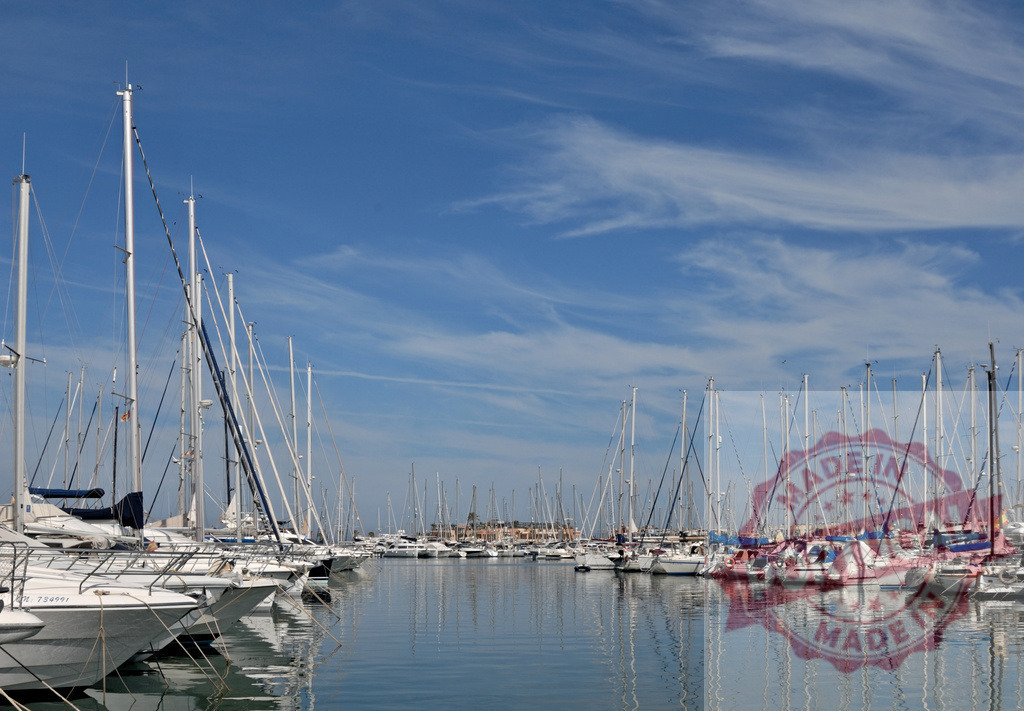 Sailboats moored in a marina reflecting in calm water | Sailboats docked in a large marina with masts reflecting on the calm blue water under a clear sky - Realized with Pictrs.com