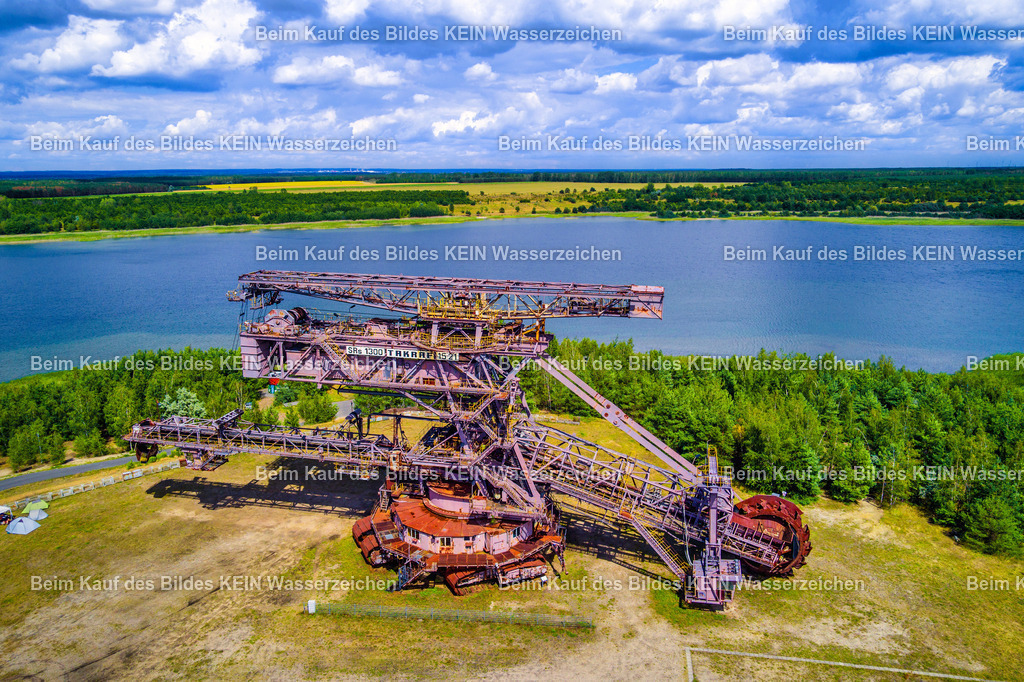Schaufelradbagger Big Wheel in Ferropolis-LU_0016 | Das Luftbild zeigt den Schaufelradbagger „Big Wheel“ in Ferropolis aus geringer Höhe. Die farbenfrohe Sommeraufnahme vermittelt eindrucksvoll die gewaltigen Dimensionen der Bergbaumaschine und macht die filigrane Stahlkonstruktion aus der Nähe besonders gut sichtbar. Aus der Drohnenperspektive werden das mächtige Schaufelrad, die weit verzweigten Förderanlagen und die markante Tragwerkskonstruktion eindrucksvoll in Szene gesetzt.Der Schaufelradbagger „Big Wheel“ wurde für den Braunkohletagebau konstruiert und gehört zu den größten erhaltenen Bergbaumaschinen Deutschlands. Die Anlage ist rund 130 Meter lang, etwa 38 Meter hoch und wiegt mehrere tausend Tonnen. Das Schaufelrad besitzt einen Durchmesser von rund 18 Metern und war in der Lage, mehrere tausend Kubikmeter Abraum pro Stunde zu fördern. Solche Maschinen arbeiteten rund um die Uhr und zählten zu den leistungsfähigsten Großgeräten des europäischen Tagebaus.Die Drohnenaufnahme dokumentiert eindrucksvoll die technische Leistungsfähigkeit und die enorme Dimension dieser historischen Bergbaumaschine. Heute ist „Big Wheel“ Bestandteil des Industriemuseums Ferropolis und erinnert als technisches Denkmal an die jahrzehntelange Braunkohleförderung in Mitteldeutschland. - Realisiert mit Pictrs.com