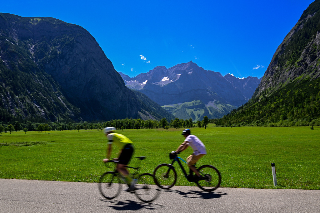 Freiheit auf zwei Rädern: Dem Horizont entgegen | Ein Anblick voller Dynamik, Lebensfreude und alpiner Freiheit: Zwei Radfahrer ziehen zügig an einer sattgrünen, weiten Ebene vorbei, getragen von der puren Lust an der Bewegung. Die leichte Unschärfe der Fahrer im Vordergrund fängt das berauschende Gefühl von Geschwindigkeit perfekt ein. Dahinter eröffnet sich die ganze, erhabene Pracht des Karwendelgebirges: Sanfte Wiesen, dichte Baumreihen und die mächtigen, felsigen Gipfel, die unter einem strahlend blauen Sommerhimmel thronen. Diese Aufnahme symbolisiert die perfekte Balance zwischen sportlicher Aktivität und der beruhigenden, zeitlosen Kraft einer majestätischen Bergwelt. - Realisiert mit Pictrs.com