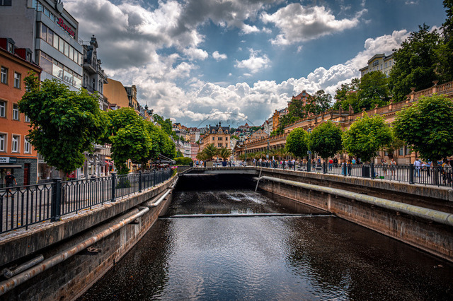 In Karlsbad fließt der Fluss ruhig durch die Stadt. Menschen gehen am Ufer entlang und genießen die Aussicht auf die schönen Gebäude und die Wolken. Es ist ein sonniger Tag | In Karlsbad fließt der Fluss ruhig durch die Stadt. Menschen gehen am Ufer entlang und genießen die Aussicht auf die schönen Gebäude und die Wolken. Es ist ein sonniger Tag. - Realisiert mit Pictrs.com