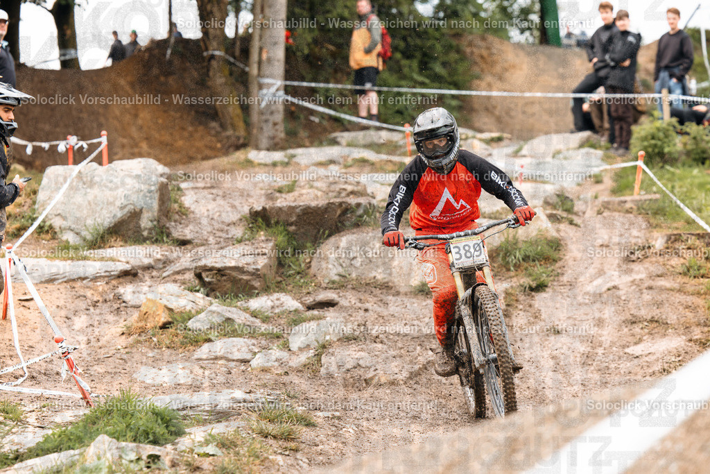 Mountainbike-Wettkampf | Ein Mountainbiker in voller Ausrüstung rast einen staubigen, felsigen Pfad hinunter. Er trägt einen schwarzen Helm mit einer orangefarbenen Weste und schwarzen Handschuhen. Sein Mountainbike hat eine Nummer 388 auf dem Rahmen. Im Hintergrund sind Zuschauer und Kameraleute zu sehen, die den Wettkampf beobachten. Der Pfad ist mit Steinen und Wurzeln übersät, was die Fahrt anspruchsvoll macht. - Realisiert mit Pictrs.com