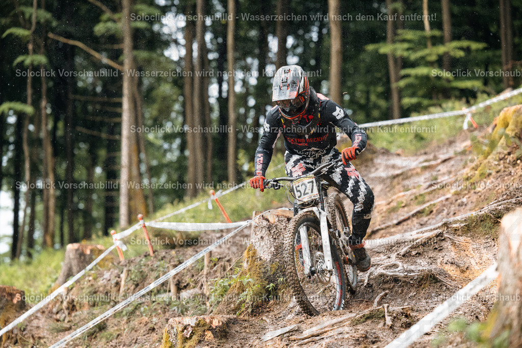 Mountainbike-Abenteuer im Wald | Das Bild zeigt einen Mountainbiker in voller Ausrüstung, der einen anspruchsvollen Trail in einem bewaldeten Gebiet hinunterfährt. Der Biker trägt einen Helm, eine Schutzbrille, Handschuhe und spezielle Kleidung, die ihn vor den Elementen und Verletzungen schützen sollen. Der Trail ist steil und schlammig, was die Fahrt zusätzlich erschwert. Im Hintergrund sind Bäume und eine Wasserfläche zu sehen. - Realisiert mit Pictrs.com