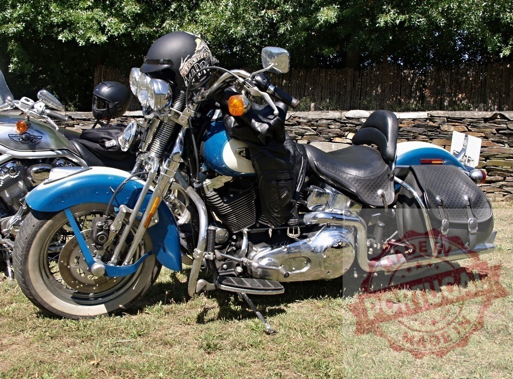 Harley-Davidson Softail Heritage Classic motorcycle parked on grass near stone wall | Parked Harley-Davidson Softail Heritage Classic motorcycle with saddlebags, crash bar, and helmet resting on handlebar - Realized with Pictrs.com