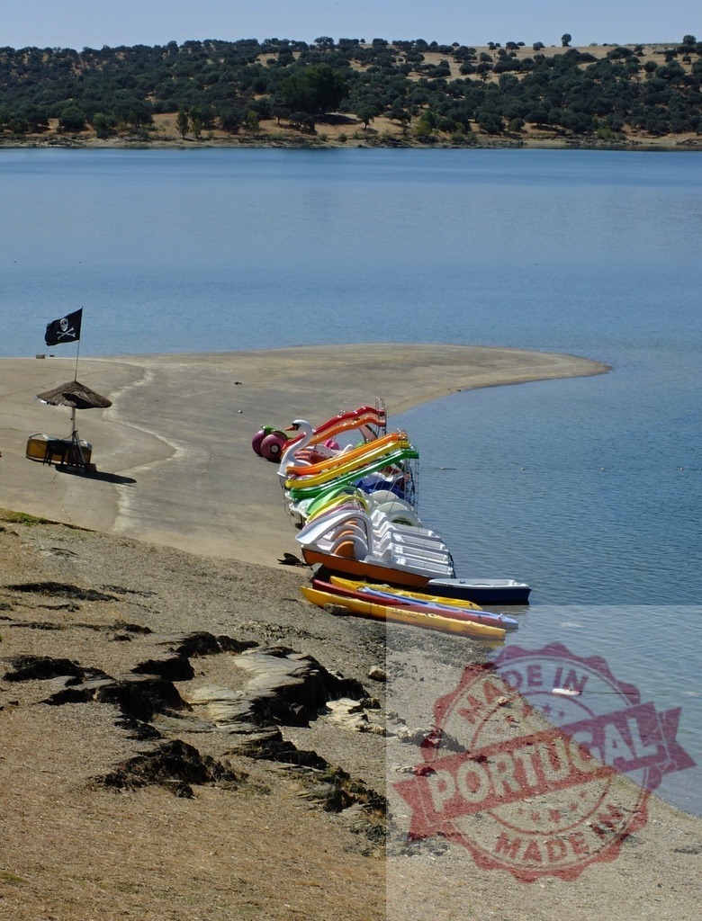 Colorful pedal boats and kayaks along sandy lake beach. Spain - Extremadura | Colorful pedal boats and kayaks parked on a sandy beach next to a tranquil blue lake under a clear sky - Realized with Pictrs.com