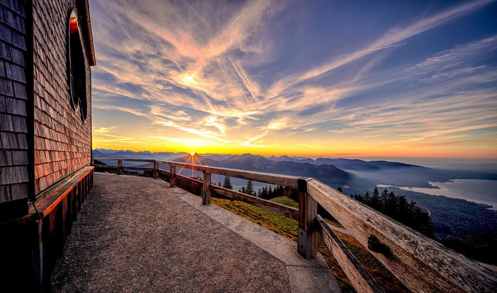 Sonnenuntergang über den Tegernsee und den bayrischen Alpen | Das Bild zeigt einen malerischen Sonnenuntergang in den Bergen, aufgenommen von der Terrasse der Kirche auf dem Wallberg. Ort: Es handelt sich höchstwahrscheinlich um die Kirche auf dem Wallberg in den Alpen.Aussicht: Der Blick geht über Bergketten, die in das Abendlicht getaucht sind, mit einem Flusslauf oder See in der Tiefe. - Realisiert mit Pictrs.com