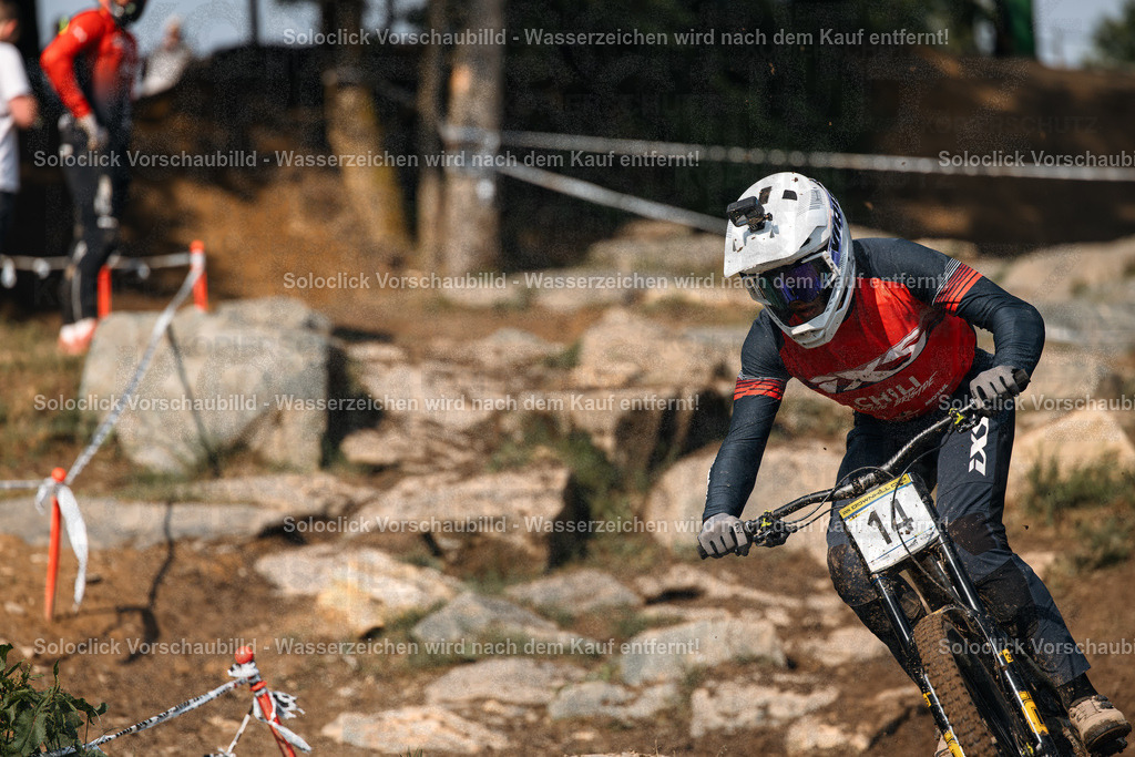 Adrenalin pur: Mountainbiker im Full Speed | Ein Mountainbiker in voller Montur rast mit hoher Geschwindigkeit einen anspruchsvollen Trail hinunter. Er trägt einen weißen Helm mit roten Akzenten, eine rote und schwarze Jacke, Handschuhe und eine Schutzbrille. Der Biker ist in Aktion und konzentriert sich voll und ganz auf die Fahrt. Der Trail ist steinig und verschlungen, mit vielen Wurzeln und Hindernissen. Es gibt rote und weiße Markierungspfähle am Wegesrand, die den Weg weisen. Im Hintergrund sind Bäume und Büsche zu sehen. - Realisiert mit Pictrs.com