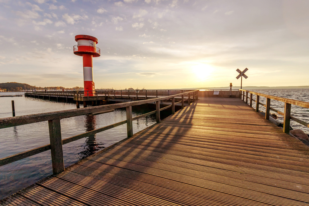 Wandbild: Bootssteg und Leuchtturm an der Ostsee | Das Wandbild im Querformat zeigt einen schönen Sonnenaufgang an der Ostsee. Der Bootssteg im Vordergrund wird durch das Licht der aufgehenden Sonne angeleuchtet. Auf der linken Seite befindet sich ein schöner rot weißer Leuchtturm.  - Realisiert mit Pictrs.com