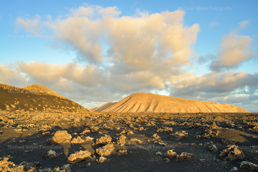 Caldera Blanca auf Lanzarote | Aus der Ebene betrachtet wirkt die Caldera Blanca wie ein mächtiger Wall, der sich aus der dunklen Lavafläche erhebt. Das Foto zeigt den Kontrast zwischen der rauen, schwarzen Landschaft im Vordergrund und den hellen Flanken des Vulkans, die sich klar gegen den Himmel abzeichnen. Von unten erscheint der Krater besonders monumental – ein stiller Zeuge der vulkanischen Vergangenheit Lanzarotes, dessen Form die Landschaft bis heute prägt. - Realisiert mit Pictrs.com