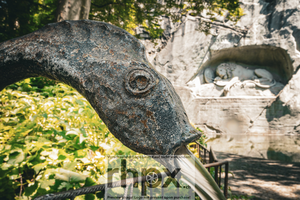 Löwendenkmal Luzern – Detail mit Brunnen / Lion Monument Lucerne – Detail with Fountain | Aufnahme des Löwendenkmals in Luzern mit einer Brunnenkulptur im Vordergrund. Das Bild zeigt den Kontrast zwischen dem bronzenen Detail und dem Felsrelief des Denkmals im Hintergrund. *************** Photograph of Lucerne’s Lion Monument with a fountain sculpture in the foreground. The image highlights the contrast between the bronze detail and the stone relief of the monument in the background. - Realisiert mit Pictrs.com