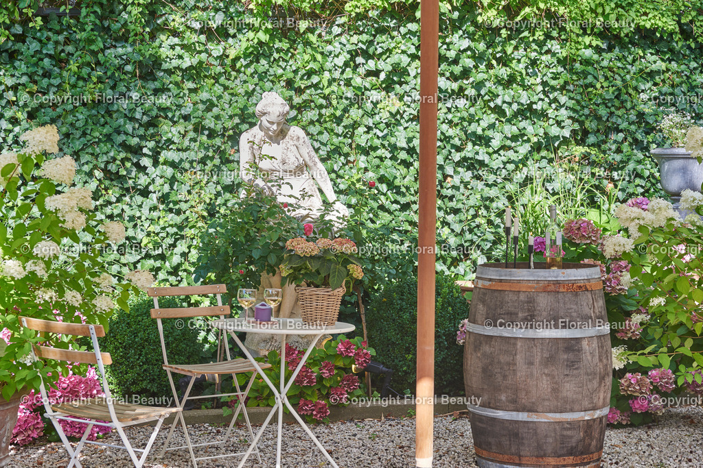 Mediterraner Stadtgarten | Gartensitzecke und Weinfass vor blühenden Hortensien mit Statue im Hintergrund - Realized with Pictrs.com