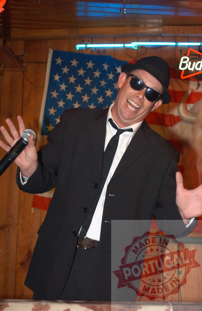 Blues Brothers impersonator singing at a bar | Man dressed as Blues Brother holding a microphone and entertaining in a bar setting - Realized with Pictrs.com