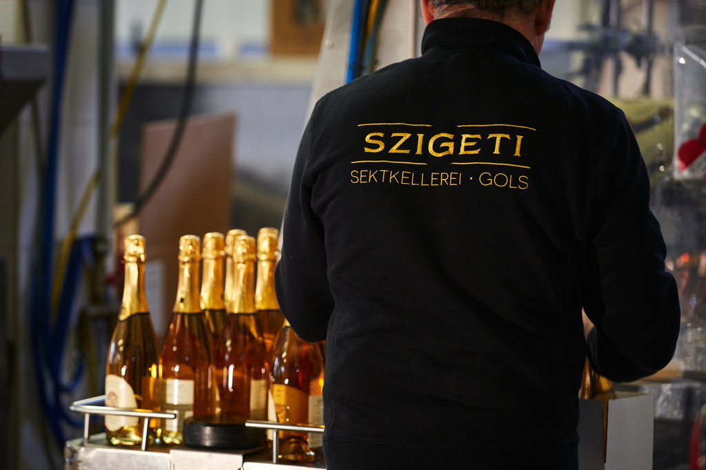 Sektkellerei Szigeti | Gols, Austria - February 19, 2018: Sektkellerei Szigeti; Mitarbeiter am Arbeitsplatz mit schwarzer Jacke und Szigeti Aufschrift, daneben die Flaschen auf dem Förderband. - Realisiert mit Pictrs.com