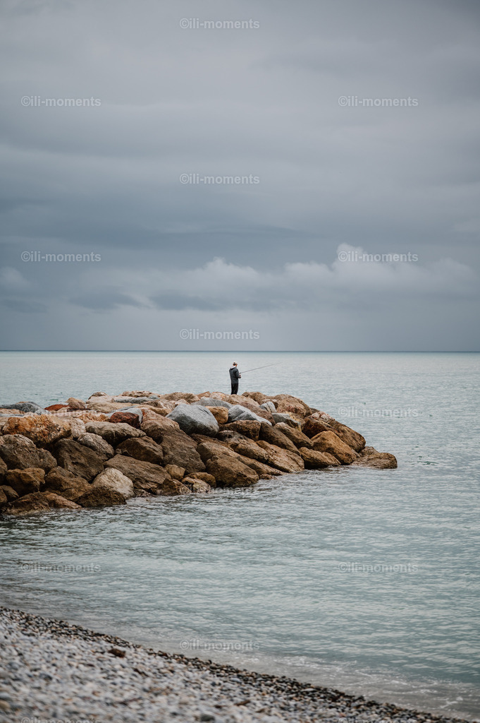 Angler am Meer | Ein Angler steht ruhig auf den Felsen am Mittelmeer und genießt die stille Atmosphäre. Dieses stimmungsvolle Foto fängt Ruhe und Gelassenheit am Meer ein – perfekt als Kunstdruck für entspannte, naturnahe Räume. - Realisiert mit Pictrs.com