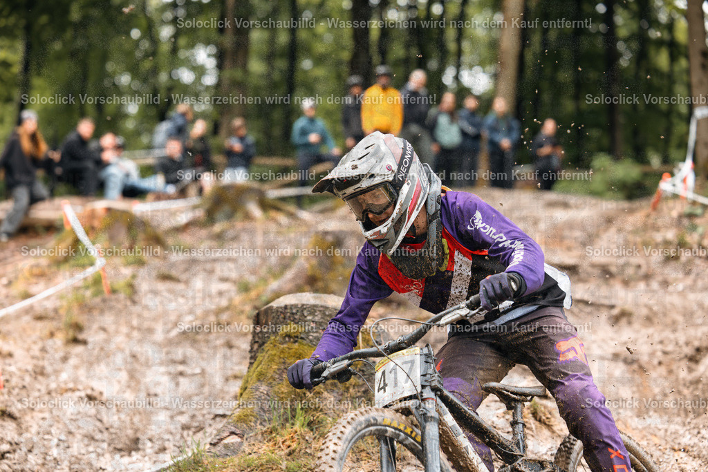 Mountainbike-Action im Wald | Das Foto zeigt einen Mountainbiker in voller Montur, der mit hoher Geschwindigkeit einen staubigen, unebenen Waldweg durchquert. Er trägt einen violetten Overall, eine schwarze Helm mit einer violetten Visier und Handschuhe. Er trägt die Nummer 417 auf seinem Fahrradrahmen. Im Hintergrund sind Zuschauer, die den Wettkampf beobachten, zu sehen. Der Wald ist dicht bewachsen und die Umgebung ist mit Wurzeln und Steinen übersät. - Realisiert mit Pictrs.com