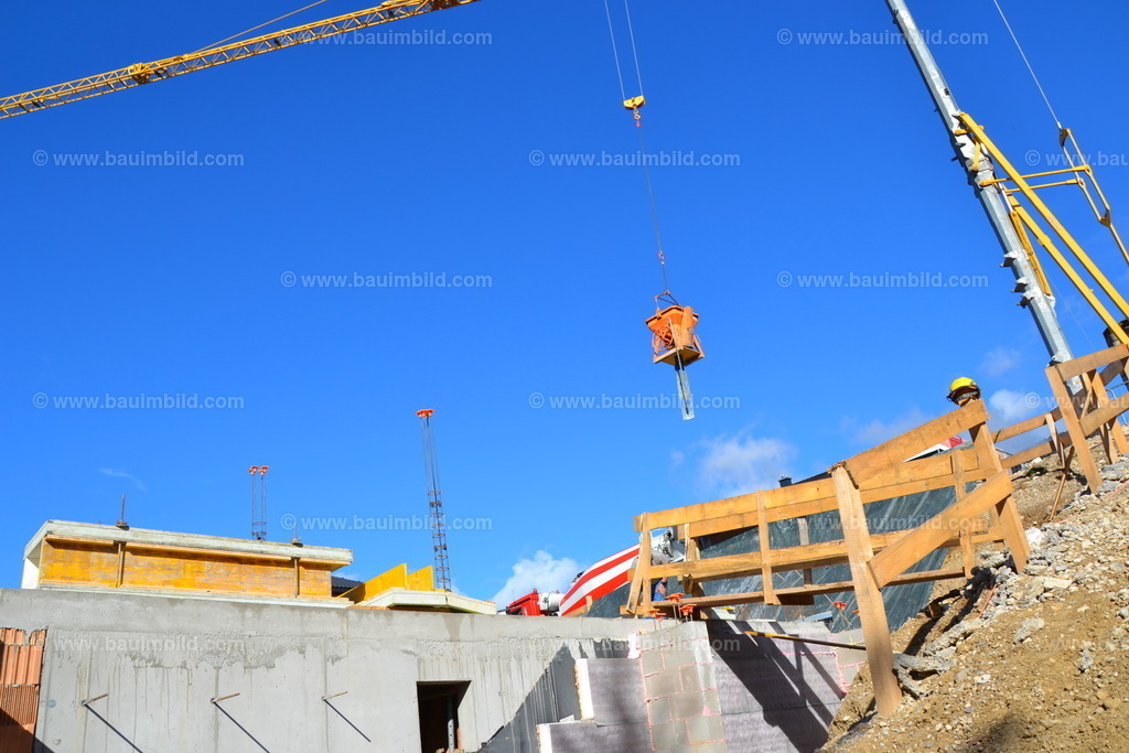 Kranarbeit | Baustelle in Hanglage mit Betonkeller und Baukran mit Krankübel, dahinter blauer Himmel