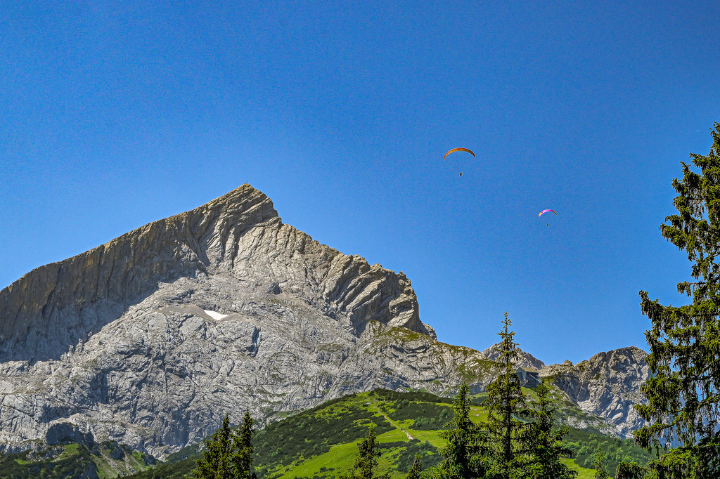 Faszination Alpen: Gipfelsturm und Freiheit | Ein atemberaubender Anblick, der die Sehnsucht nach den Bergen weckt: Majestätisch und unbezwingbar ragt die markante Pyramidengestalt der Alpspitze in den strahlend blauen Himmel empor. Ihre steilen Felswände und bizarren Grate erzählen von jahrtausendealter Erdgeschichte und der rohen Kraft der Natur. Zwei Gleitschirmflieger, winzige Punkte vor der gewaltigen Kulisse, unterstreichen die Dimensionen und die unendliche Weite der Bergwelt. Dieses Bild ist eine Hommage an die Schönheit der Alpen und den Traum von Freiheit und Abenteuer. - Realisiert mit Pictrs.com