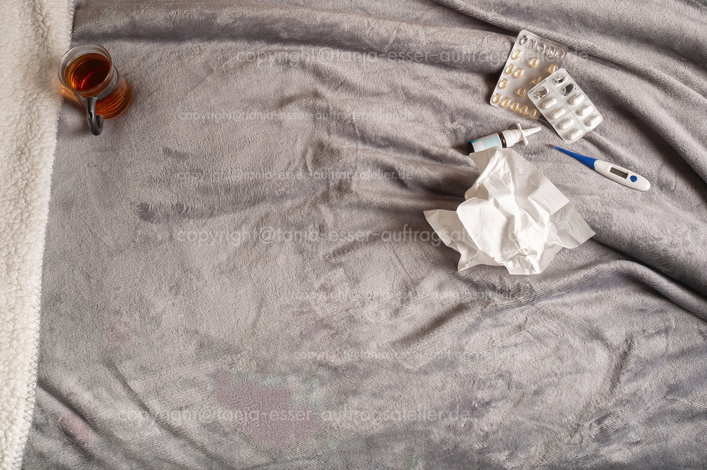 Erkältungstee D | Auf einer kuscheligen grauen Decke mit Sherpa Fell sind verschiedene Utensilien zur Behandlung eines Erkältungsinfekts arrangiert. Ein Glas heißer Tee, Taschentücher, ein Fieberthermometer, Nasenspray und mehrere Blister mit Medikamenten 