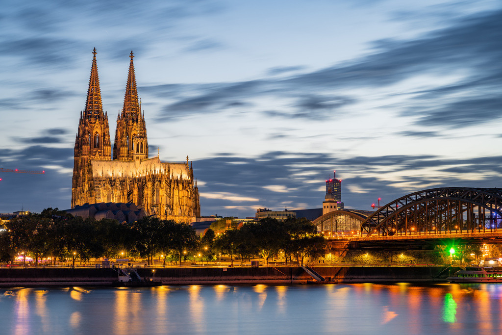 Kölner Dom und Rheinpanorama zur blauen Stunde | Der beleuchtete Kölner Dom und die Hohenzollernbrücke prägen das abendliche Rheinpanorama. Die Spiegelungen im Wasser und die ziehenden Wolken verleihen der klassischen Stadtansicht eine ruhige, elegante Wirkung. - Realisiert mit Pictrs.com