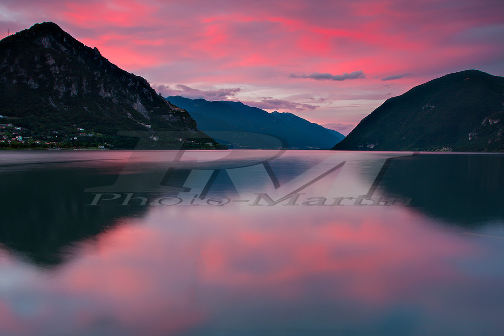 Lago d’Idro bei Sonnenuntergang | Der Lago d’Idro zeigt sich bei Sonnenuntergang in ruhiger Stimmung, mit rosa Himmel, spiegelglattem Wasser und majestätischer Bergkulisse. - Realisiert mit Pictrs.com