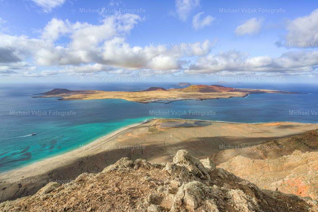 Blick von Lanzarote nach La Graciosa | Etwas südlich des Mirador del Río öffnet sich der Blick über das Meer zur Insel La Graciosa. Die kargen Klippen Lanzarotes rahmen die Szene, während die kleine Nachbarinsel wie ein heller Streifen im tiefen Blau des Atlantiks liegt. Das Licht betont die goldbraunen Flächen und zeichnet klare Konturen zwischen Wasser, Himmel und vulkanischem Gestein. - Realisiert mit Pictrs.com