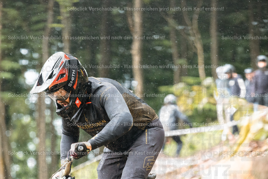 Mountainbiker im Regen | Das Bild zeigt einen Mountainbiker in voller Montur, der einen steilen, mit Erde und Schlamm bedeckten Hang hinunterfährt. Er trägt einen schwarzen Helm mit der Aufschrift 'SMITH' und eine dunkle Weste mit dem Schriftzug 'GLERSTUBEN'. Der Hintergrund zeigt einen bewaldeten Hang mit anderen Fahrern und Zuschauern, die im Regen stehen. Das Wetter ist regnerisch und die Atmosphäre ist dynamisch und herausfordernd. - Realisiert mit Pictrs.com