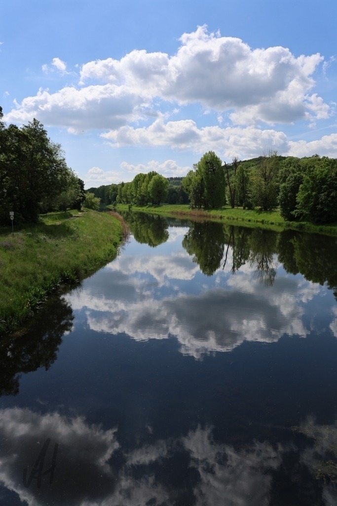 Wolkenspiegelung im Fluss der Weißen Elster | Die Aufnahme zeigt die ruhige Oberfläche der Weißen Elster am Wehr von Bad Köstritz, in der sich ein dramatischer Himmel mit großen, weichen Wolken spiegelt. Durch das fast unbewegte Wasser wirkt die Spiegelung beinahe wie ein zweites Bild unterhalb des Horizonts. Die Ufer des Flusses rahmen die Szene natürlich ein – mit Bäumen und Wiesen im Hintergrund – und verleihen dem Motiv Tiefe.Das Licht ist sanft und diffus. Dadurch entstehen feine Kontraste zwischen Himmel, Wasser und Landschaft. Die Wolken wirken plastisch und ziehen den Blick in die Spiegelung hinein, sodass Realität und Reflexion fast miteinander verschmelzen.Die Stimmung des Fotos ist still, meditativ und naturverbunden. Besonders die Kombination aus Himmel und Wasser vermittelt Weite und Gelassenheit. - Realisiert mit Pictrs.com