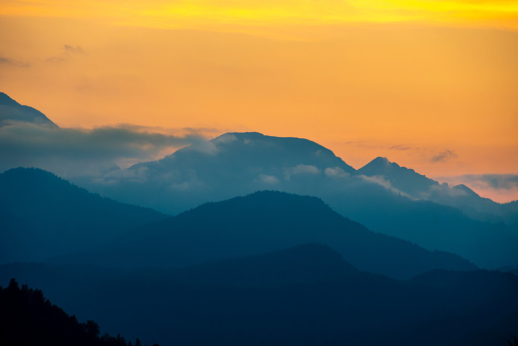 Goldenes Abendglühen über den Alpen | Sanft gestaffelte Bergkulissen zeichnen sich in kühlen Blautönen vor einem magisch leuchtenden, orange-goldenen Abendhimmel ab. Zarte Nebelschleier schmiegen sich an die Hänge der Voralpenkette nahe Garmisch-Partenkirchen und verleihen der Szenerie eine geheimnisvolle Tiefe. Dieses stimmungsvolle Farbenspiel fängt die friedvolle Stille eines ausklingenden Sommertages in den Bergen ein – ein erhabener Moment der Ruhe, der zum Träumen und Innehalten einlädt. - Realisiert mit Pictrs.com