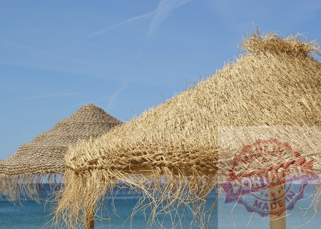 Straw beach umbrellas protecting from sun on tropical coastline | Straw sunshades providing shade on a sandy beach by the clear blue sea - Realisiert mit Pictrs.com