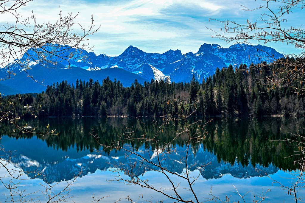 Spiegelbild der Ewigkeit: Magische Stille am Barmsee | in Anblick von vollkommener Harmonie und tiefer, innerer Ruhe: Wie ein riesiger, gläserner Spiegel wirft der tiefblaue Barmsee zwischen Garmisch-Partenkirchen und Mittenwald das majestätische Panorama der schneebedeckten Karwendel-Gipfel zurück. Die dichten Nadelwälder am Ufer trennen Himmel und Wasser mit einer Linie aus sattem Dunkelgrün, während zarte, noch blattlose Frühlingszweige im Vordergrund das Bild wie ein natürlicher Rahmen umgeben. Diese Aufnahme fängt die absolute Windstille und die erhabene Klarheit der Natur perfekt ein – ein magischer Moment der Entschleunigung, der die Seele tief durchatmen lässt. - Realisiert mit Pictrs.com