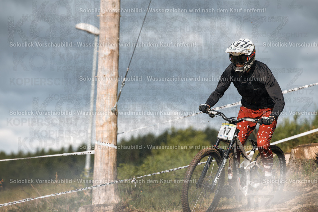 Mountainbike-Action | Ein Mountainbiker rast auf einem staubigen Pfad hinab, während er einen Wettkampf bestreitet. Er trägt einen schwarzen Overall, eine weiße und schwarze Helm und Handschuhe. Der Hintergrund besteht aus Bäumen und einem bewölkten Himmel. Staub wirbelt um seine Reifen auf. - Realisiert mit Pictrs.com