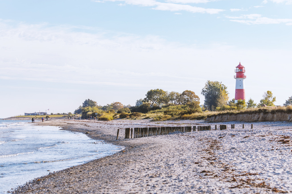 Wandbild: Küstenlinie Falshöft - Leuchtturm am Strand | Dieses charakteristische Wandbild fängt den zeitlosen Charme der Ostseeküste in seiner ganzen Vielfalt ein. Der natürliche Strandverlauf mit den hölzernen Buhnen leitet den Blick sanft am Wasser entlang, während der rot-weiße Leuchtturm Falshöft harmonisch in die Landschaft eingebettet ist. Die warmen, natürlichen Töne des Sandes und die sanfte Gischt verleihen Räumen eine behagliche, elegante Atmosphäre – eine stilvolle Hommage an den Norden für anspruchsvolle Wohnkonzepte.