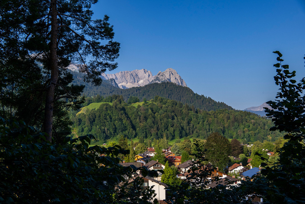 Majestätische Gipfel: Blick auf Zugspitze und Waxensteine | Durch einen natürlichen Rahmen aus schattenspendendem Laub blickt man auf die idyllischen Dächer von Garmisch-Partenkirchen. Dahinter steigen dicht bewaldete Hänge empor, über denen sich die sonnenbeschienenen, mächtigen Massive der Zugspitze und der kühnen Waxensteine im tiefblauen Sommerhimmel abzeichnen. Die Komposition verbindet das geborgene Gefühl eines gemütlichen Bergdorfs mit der imponierenden Weite der Alpen. Eine Aufnahme, die Ruhe, Naturverbundenheit und bayerische Lebensfreude ausstrahlt. - Realisiert mit Pictrs.com