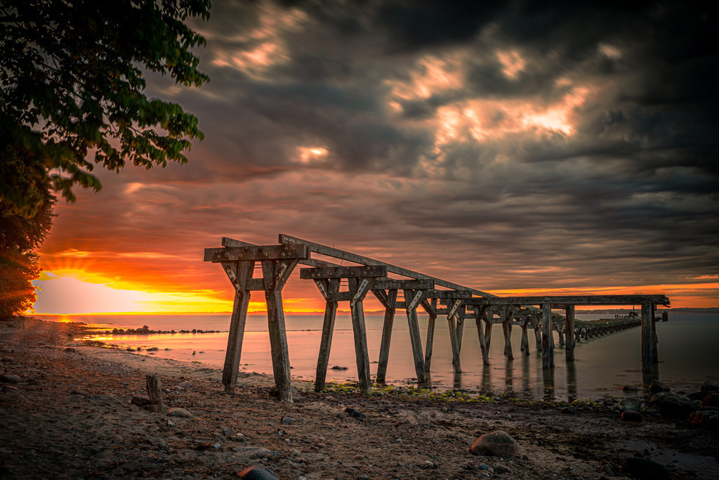 Alte Seebrücke von Quern an der Flensburger Förde | Auf dem Foto sind die Überreste der Beeindruckenden alte Seebrücke von Quern an der Flensburger Förde zu sehen. Die robusten Holzpfähle zeigen Spuren vergangener Tage. Das Foto fängt die nostalgische Schönheit und den Charme der alten Seebrücke von Quern ein. Es erinnert an vergangene Zeiten, als die Seebrücke noch ein belebter Treffpunkt für Einheimische und Touristen war. Gleichzeitig strahlt es Ruhe und Gelassenheit aus und lädt den Betrachter ein, einen Moment der Stille und des Innehaltens zu genießen.