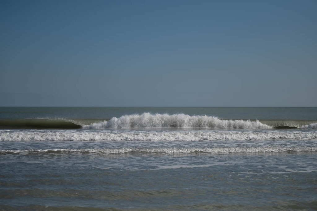 Küstenaufnahme von Lido Salpi, Manfredonia | Die Aufnahme zeigt eine Uferlinie des Meeres mit wellen, die an den Strand rollen. Der Himmel ist blau und die Küstenlinie ist mit etwas Vegetation übersät. Die Koordinate des Aufnahmeortes sind 41.555131814397, 15.893422331352. Der Kontext deutet darauf hin, dass das Foto am Strand des Camping Lido Salpi in Manfredonia (Foggia) aufgenommen wurde. - Realisiert mit Pictrs.com