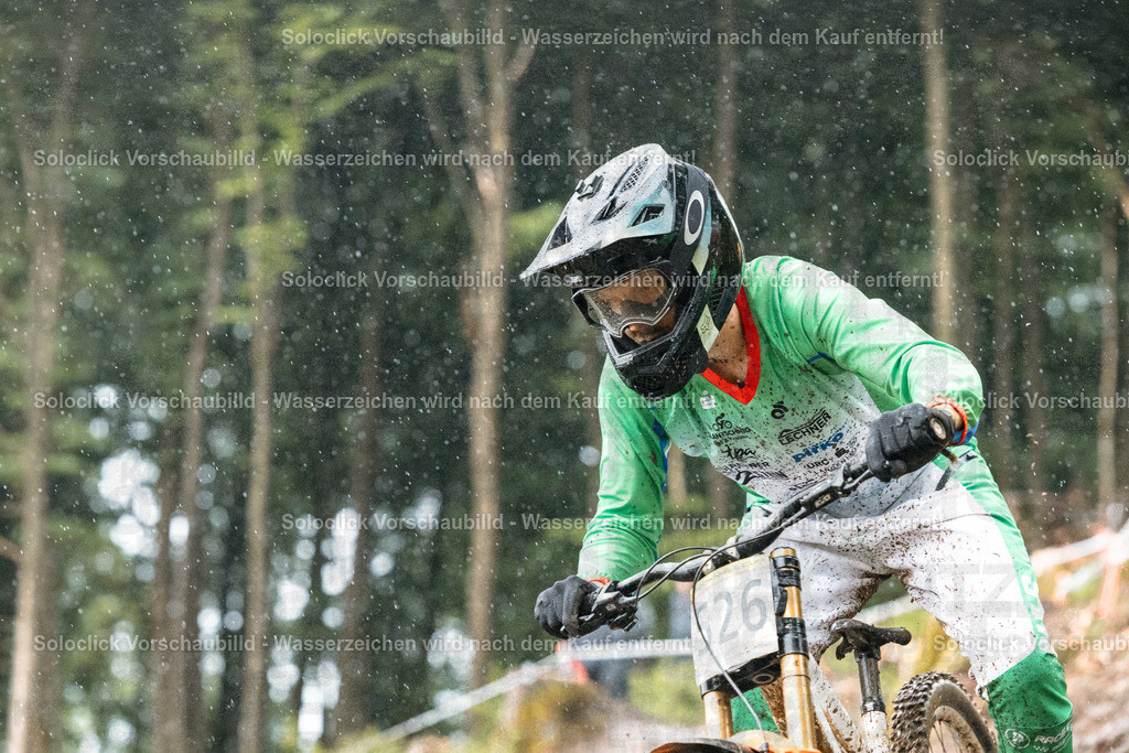 Mountainbike-Action im Regen | Ein Mountainbiker in voller Ausrüstung rast durch einen bewaldeten Hang. Er trägt einen grünen Trikot, eine schwarze Hose und einen schwarzen Helm mit grünen Akzenten. Sein Gesicht ist durch eine dunkle Maske verdeckt, und er blickt konzentriert auf die Strecke vor ihm. Der Hintergrund besteht aus dichtem Grünwerk, das von Regen benetzt ist. Der Fahrer trägt die Nummer 26 auf seinem Rücken. - Realisiert mit Pictrs.com