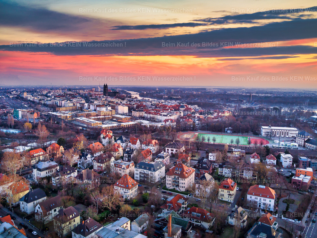 Magdeburg_Luftbild_Lenneviertel_Jean-Burger_Erich-Weinert_Morgenrot_AMO-0021 | Dieses Luftbild zeigt das Magdeburger Lenneviertel in den frühen Morgenstunden eines farbenprächtigen Sonnenaufgangs. Die aufgehende Sonne taucht den Himmel über der Landeshauptstadt in intensive Rot-, Orange- und Violetttöne. Durch die stimmungsvolle Lichtstimmung und die gezielte Bildbearbeitung entsteht ein besonders eindrucksvoller Blick auf eines der charakteristischsten Wohnviertel Magdeburgs.Im Mittelpunkt der Aufnahme liegt das historische Lenneviertel zwischen Jean-Burger-Straße und Humboldtstraße. Die gründerzeitliche Bebauung mit ihren geschlossenen Straßenzügen, Innenhöfen und markanten Dachlandschaften kommt aus der Vogelperspektive besonders gut zur Geltung. Die unbelaubten Bäume unterstreichen die winterliche Jahreszeit und ermöglichen zugleich einen freien Blick auf die architektonischen Strukturen des Viertels.Aus der Luft wird sichtbar, wie harmonisch sich das Lenneviertel in das Stadtbild Magdeburgs einfügt. Die Kombination aus historischer Architektur, klaren Linien der Straßenführung und dem spektakulären Morgenhimmel verleiht dieser Luftaufnahme eine ganz besondere Atmosphäre und macht sie zu einem eindrucksvollen Dokument der Stadtentwicklung Magdeburgs. - Realisiert mit Pictrs.com