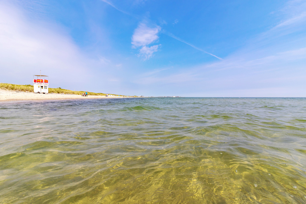 Wandbild: Blick vom Meer zum Strand in Schönhagen | Dieses Wandbild im Querformat zeigt den Strand in Schönhagen vom Meer aus. Das Wasser ist sehr klar. Am blauen Himmel befinden sich nur einige Schleierwolken. - Realisiert mit Pictrs.com