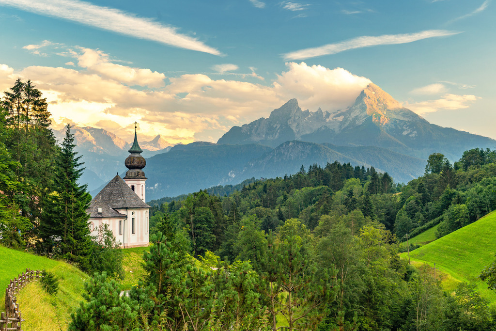 Wandbild: Idyllische Alpenlandschaft: Maria Gern und der Watzmann | Dieses wunderschöne Bild zeigt die malerische Kapelle Maria Gern in Bayern, eingebettet in die sanfte Hügellandschaft vor dem imposanten Hintergrund des Watzmanns, eines der bekanntesten Berge der bayerischen Alpen. Die spätbarocke Kirche mit ihrem charakteristischen Zwiebelturm steht auf einer grünen Anhöhe, umgeben von dichtem Wald und saftigen Wiesen. Der Himmel ist klar, nur von einigen dekorativen Wolken durchzogen, die in der Abendsonne leuchten und eine friedliche, fast märchenhafte Atmosphäre schaffen. Die landschaftliche Schönheit und die ruhige Erhabenheit der Alpen spiegeln sich perfekt in dieser idyllischen Szene wider.