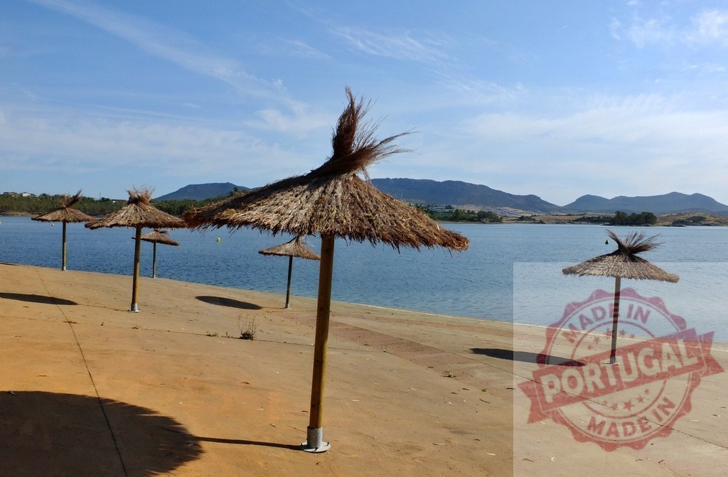 Straw umbrellas standing by lake with mountains background. Spain - Extremadura | Straw umbrellas providing shade on the shoreline of a tranquil lake under a clear blue sky - Realized with Pictrs.com