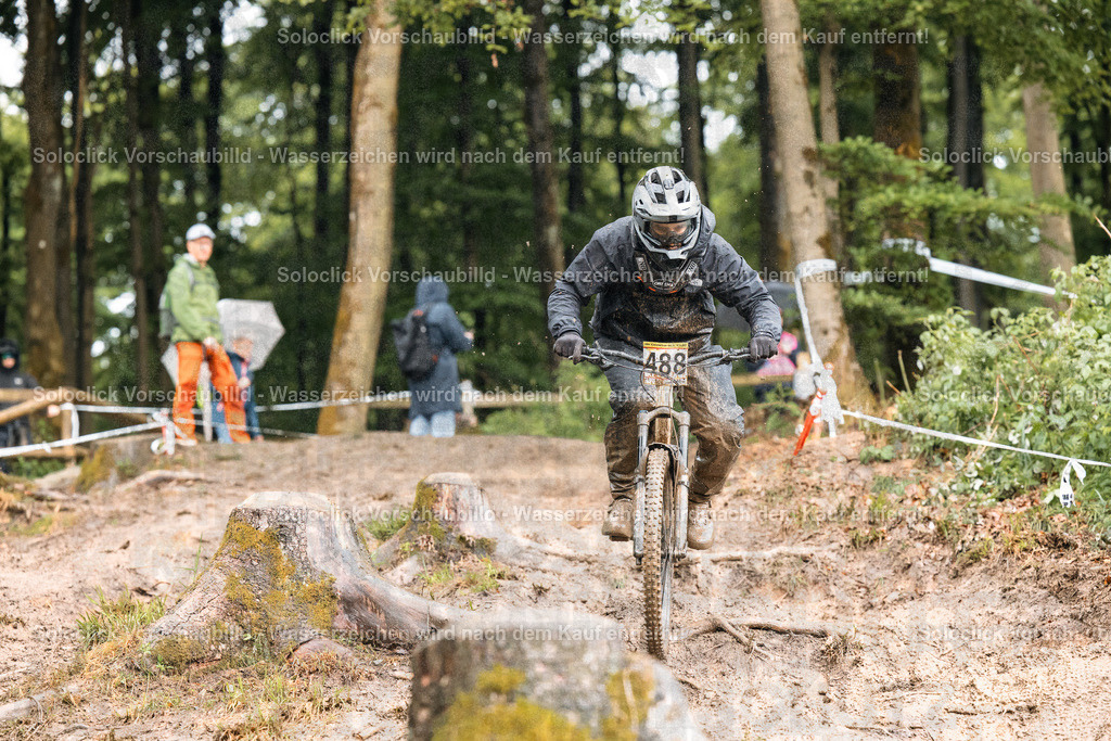 Mountainbike-Abenteuer im Wald | Ein Mountainbiker in voller Ausrüstung rast auf einem staubigen, unebenen Pfad durch einen dichten Wald. Er trägt einen schwarzen Helm und eine schwarze Maske, die sein Gesicht bedecken. Auf seinem Rücken befindet sich eine Nummer 488. Der Pfad schlängelt sich zwischen Baumstümpfen und Wurzeln hindurch. Im Hintergrund sind weitere Personen in Sportkleidung zu sehen, die vermutlich Zuschauer oder andere Teilnehmer sind. Die Szene vermittelt ein Gefühl von Geschwindigkeit und Abenteuer. - Realisiert mit Pictrs.com