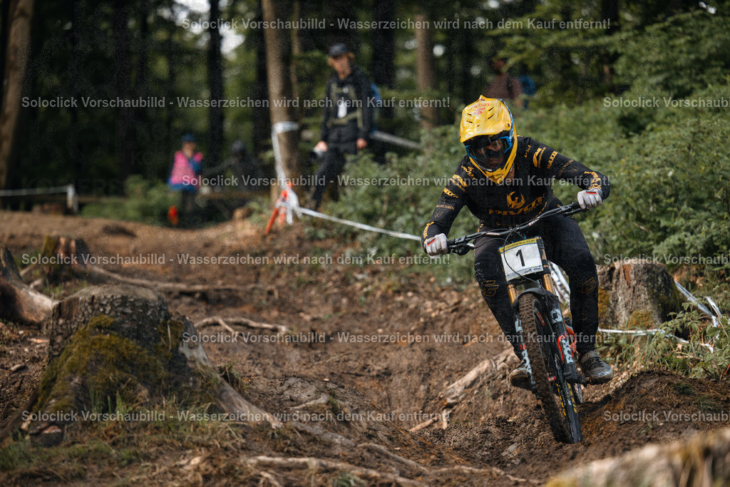 Adrenalin pur: Mountainbiker im vollen Einsatz | Ein Mountainbiker rast mit hoher Geschwindigkeit einen steilen, verschneiten Trail hinunter. Er ist in voller Montainbike-Ausrüstung gekleidet und konzentriert sich voll und ganz auf die bevorstehende Herausforderung. Der Fahrer ist mit einem gelben Helm und einer gelben Jacke ausgestattet und trägt ein Nummernschild mit der Nummer 1. Der Hintergrund zeigt einen dichten Wald, der von einem Team von Zuschauern gesäumt ist, die den Fahrer aufmerksam beobachten. - Realisiert mit Pictrs.com