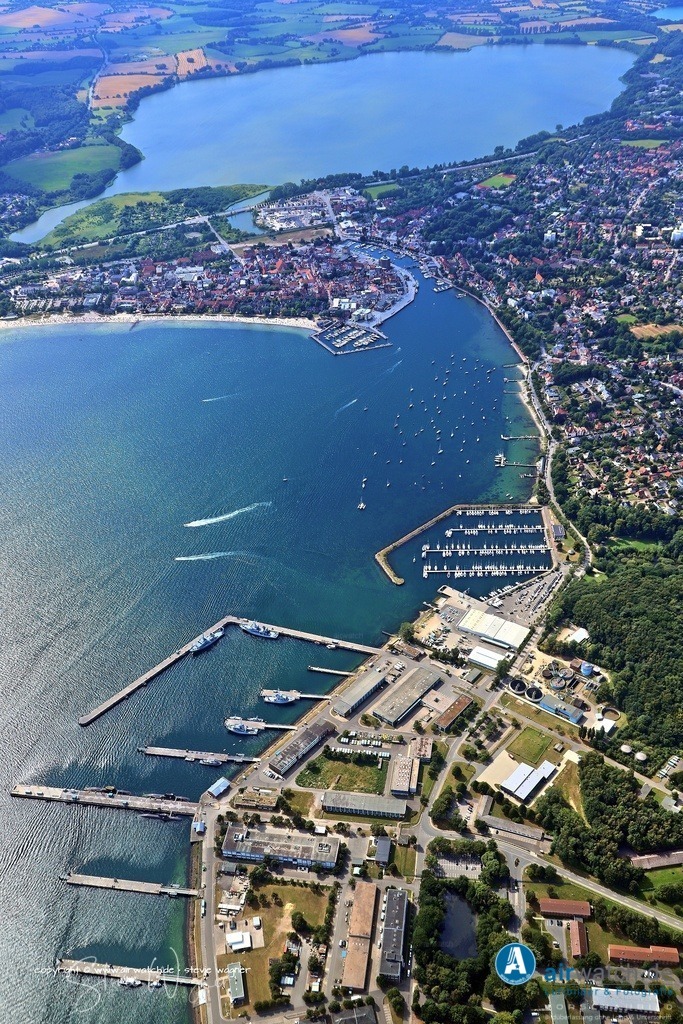Luftbild Eckernförde - Die Geschichte des Marinestützpunkts Eckernförde | Die Geschichte des Marinestützpunkts Eckernförde reicht weit zurück und ist tief in der deutschen Militärgeschichte verwurzelt. Bereits am 9. Juni 1913 wurde in Eckernförde ein Schießstand für Torpedos der Kaiserlichen Marine eingeweiht, der als Außenstelle der „Torpedowerkstatt Kiel-Friedrichsort“ diente. Die natürliche Tiefe der Eckernförder Bucht und ihre geschützte Lage machten sie zu einem idealen Standort für die Erprobung von Torpedos. Während des Ersten Weltkriegs wuchs die Bedeutung der Anlage erheblich, und bis 1918 waren 6.000 Menschen am Sandkrug beschäftigt. Nach dem Versailler Vertrag wurde die Kaiserliche Torpedowerkstatt aufgelöst, doch der Schießstand in Eckernförde blieb erhalten und wurde 1919 unter dem Namen „Torpedoversuchsanstalt“ (TVA) wieder aktiviert. In der Zeit des Nationalsozialismus wurde die Anlage erneut stark ausgebaut, und die Zahl der Beschäftigten stieg rasant an – bei Kriegsbeginn 1939 arbeiteten 3.200 Menschen in der TVA. Mit dem Kriegsende 1945 wurde die TVA stillgelegt und schließlich zwischen Dezember 1948 und Januar 1950 durch britische Besatzungstruppen abgerissen.